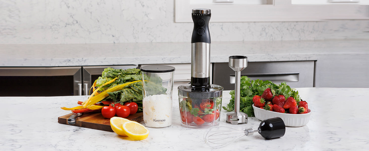 Kenmore hand blender and attachments surrounded by vegetables, greens, fruit, vegetables, and berries on a marbled white kitchen counter