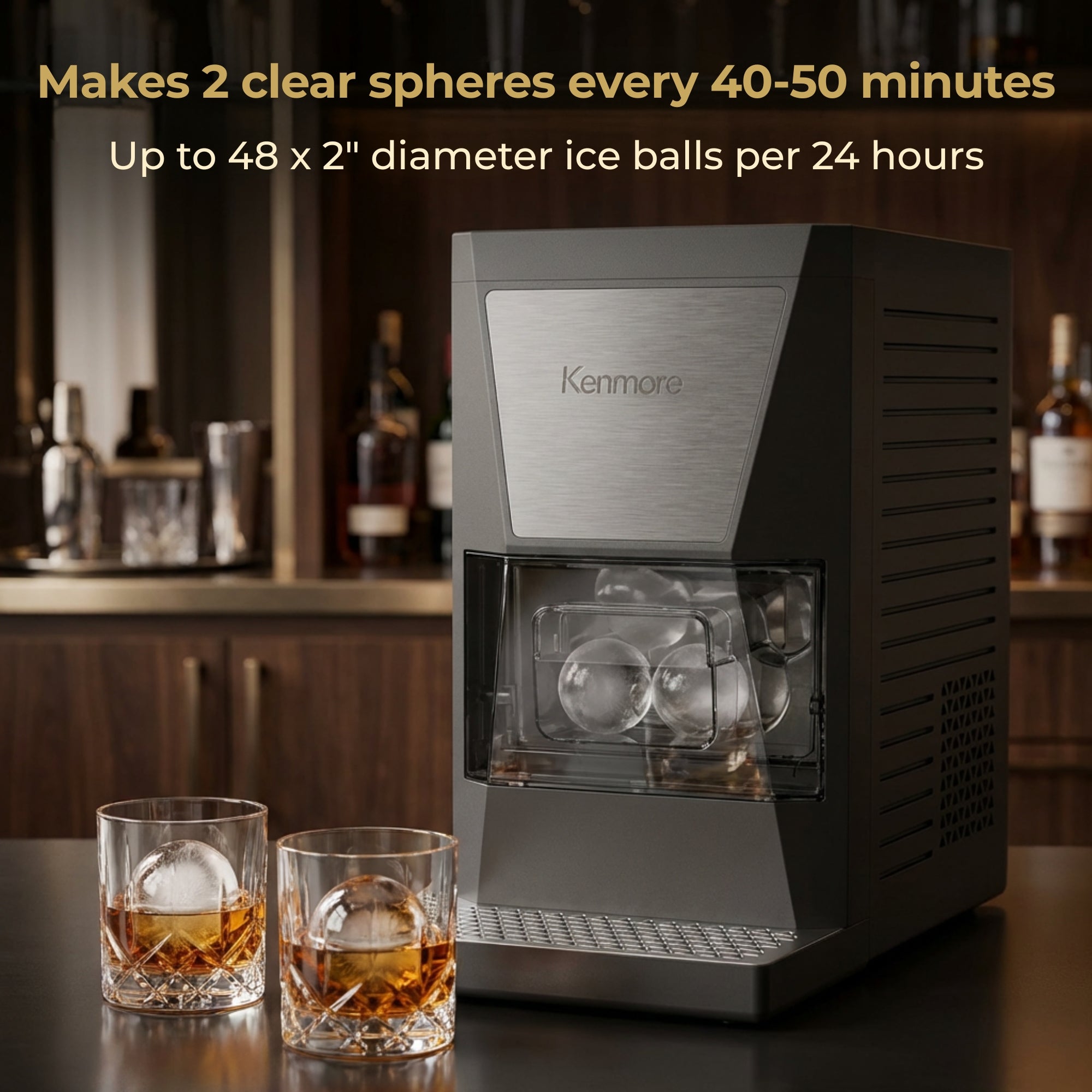 Kenmore clear sphere ice maker filled with large ice balls on a wooden countertop with two glasses containing amber-colored liquid and large round ice cubes and a bar setting in the background. Text above reads, "Makes 2 clear spheres every 40-50 minutes; Up to 48 x 2" diameter ice balls per 24 hours"