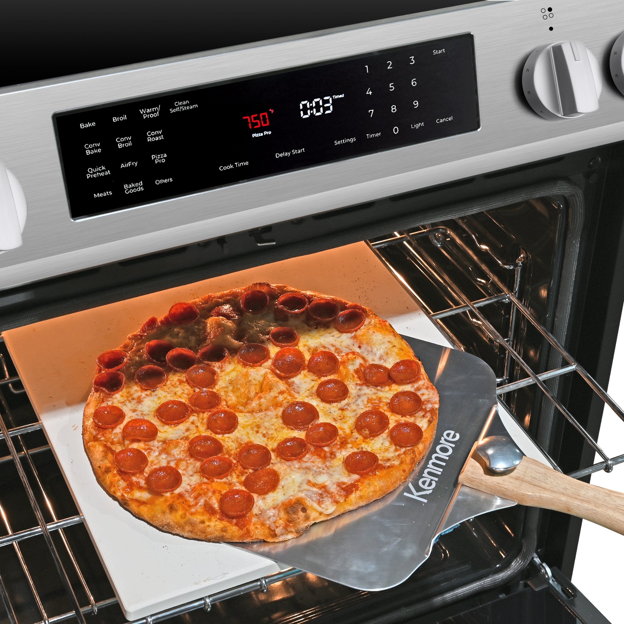 Closeup showing the control panel and top portion of the interior of the open Kenmore Pizza Pro 5.6 cu ft induction range. There is a pizza peel being used to lift a cooked pepperoni pizza from the pizza stone on the top rack of the oven.