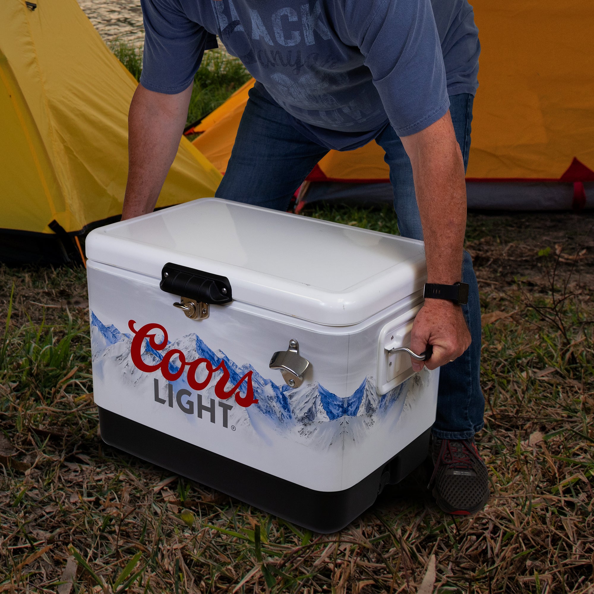 Lifestyle image of a person wearing jeans and a blue t-shirt lifting the Coors Light 54 quart ice chest with two yellow dome tents visible in the background