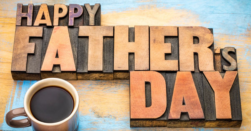 Picture shows a 3 dimensional wooden carving of the words Happy Father's Day on a blue and tan wood surface with a mug of black coffee