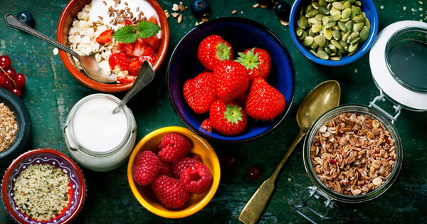 Photo shows bowls and jars of plain yogurt, yogurt topped with berries and oats, and bowls and jars of berries, seeds, and granola, on a dark green tabletop