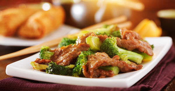 Photo of beef and broccoli on a white plate with a pair of wooden chopsticks in the background