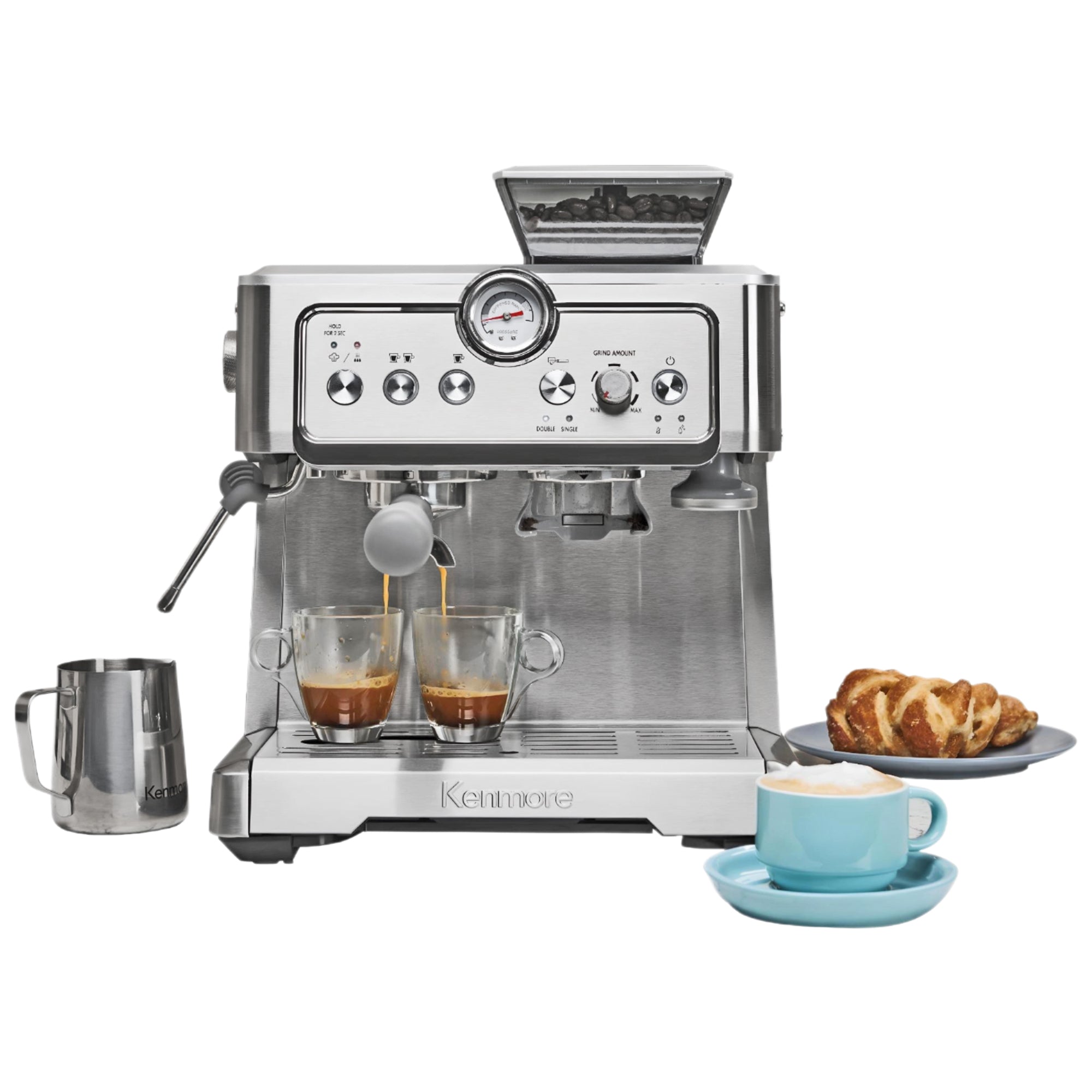 Kenmore semi-automatic espresso maker with espresso being extracted into two clear cups and a stainless steel milk pitcher, a pastry, and a cappuccino in a light blue mug arranged around it on a white background