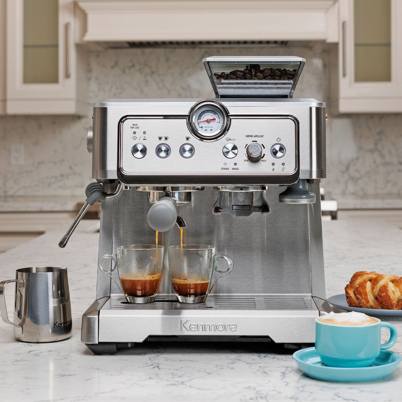 Kenmore semi-automatic espresso maker with espresso being extracted into two clear cups on a white marble countertop with the milk pitcher, a pastry, and a cappuccino in a light blue mug arranged around it.