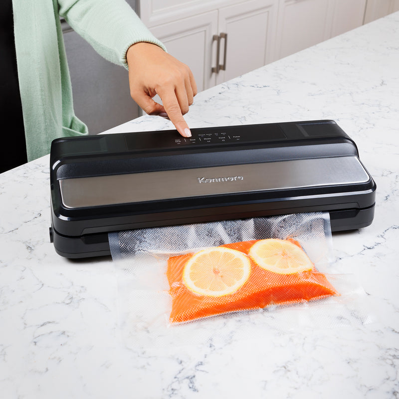 Kenmore vacuum sealer on a white and gray marble surface with a bag containing salmon and lemon slices and a person in a pale green cardigan touching the controls