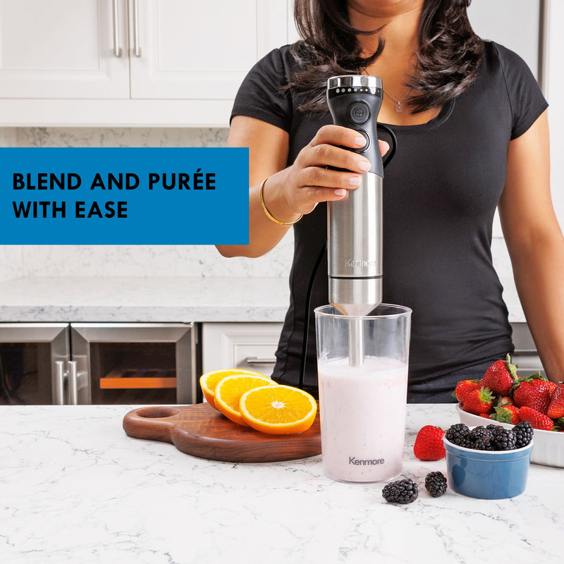 Light-skinned person in an olive-green shirt using the hand blender to puree an orange-coloured smoothie on a beige counter. There are oranges and peaches beside a glass of orange smoothie in the foreground and another kitchen counter with bowls of fruit in the background. Text overlay reads Blend and puree with ease. 