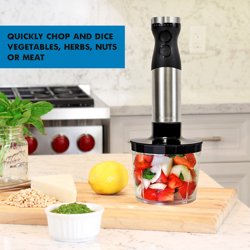 Immersion blender with food chopper attachment filled with cut up peppers, tomatoes, onions, and cilantro on a wooden cutting board with basil, pinenuts, cheese, a lemon, and a small dish of pesto beside it. Text overlay reads Quickly chop and dice vegetables, herbs, nuts, or meat.