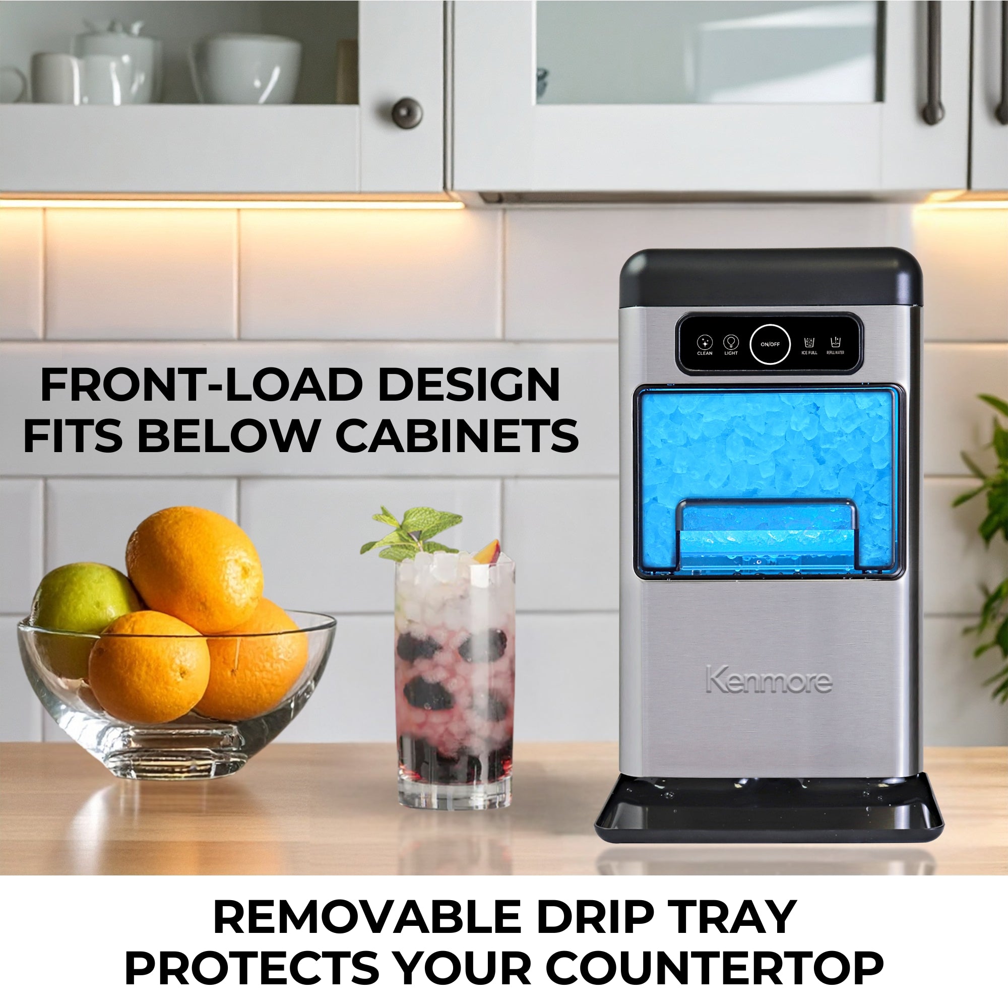 Kenmore stainless steel chewable ice maker on a wooden counter with white cupboards above it and a bowl of oranges and glass of nugget ice beside. Text to the left reads, "Front-load design fits below cabinets," and text below reads, "Removable drip tray protects your countertop"