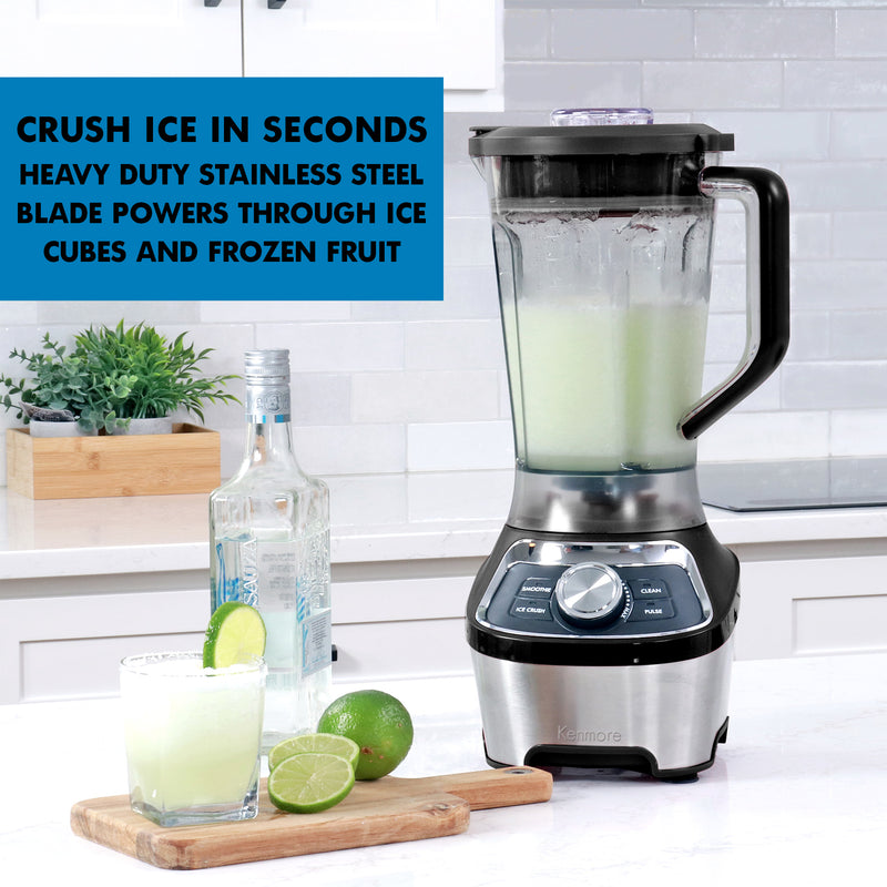 1.9 liter capacity blender filled with a light green frozen beverage on a white kitchen counter with a clear glass bottle and sliced lime and  a glass of frozen blended margarita with a salted rim on a wooden cutting board beside it. Text overlay reads Crush ice in seconds: Heavy duty stainless steel blade powers through ice cubes and frozen fruit.