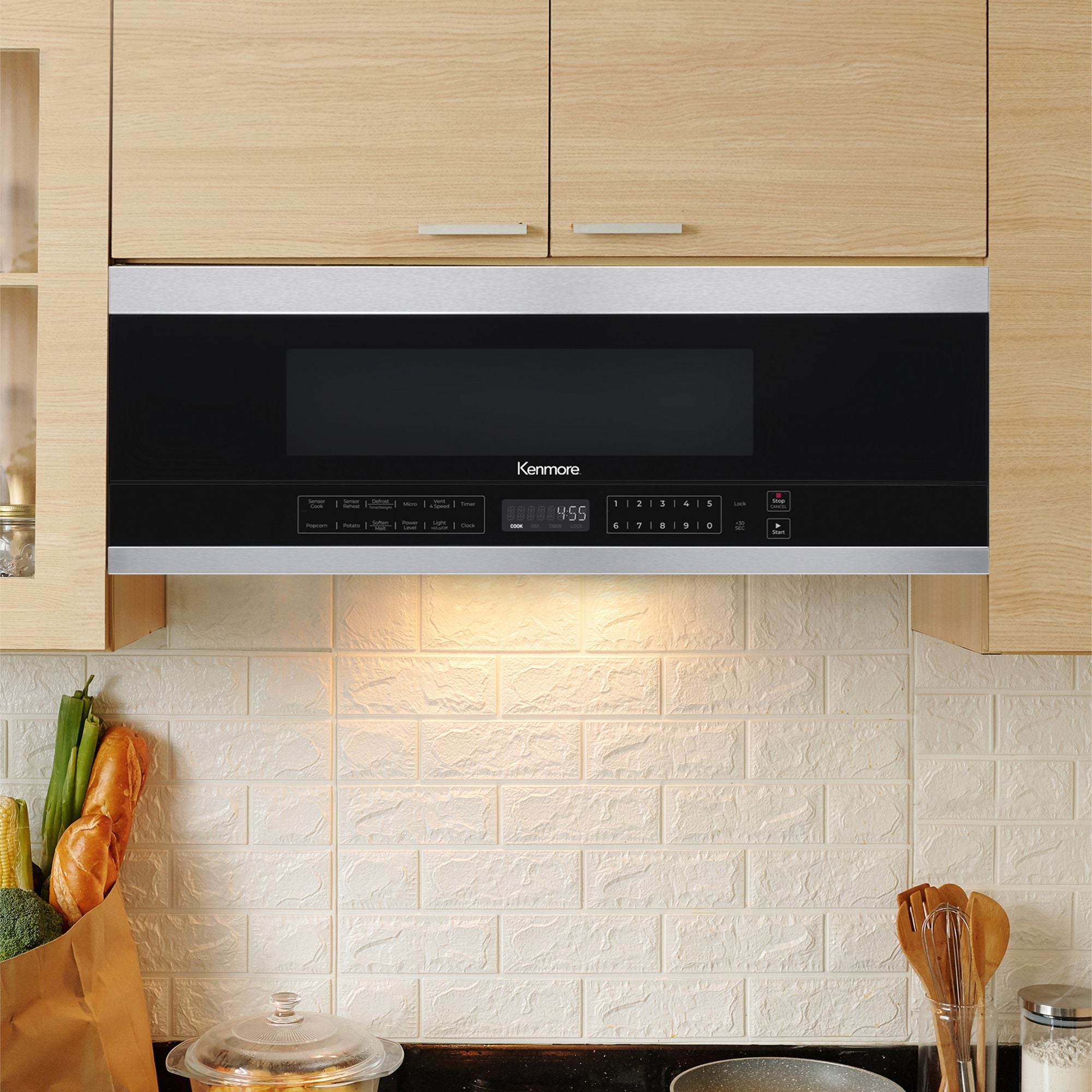 Kenmore over-the-range microwave installed between beige cupboards with the under-cabinet light on and a white tile backsplash behind.