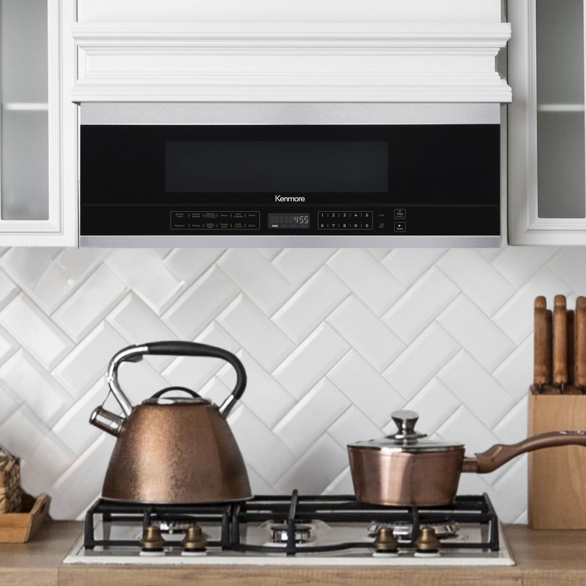 Kenmore over-the-range microwave installed between white cupboards. There is a gas range below with a copper kettle and pot on it.