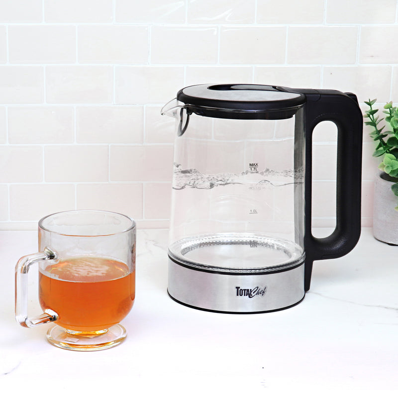 Total Chef cordless pouring electric kettle filled with boiling water with a clear glass mug of orange-colored tea beside it on a white marble countertop in front of a white tile backsplash