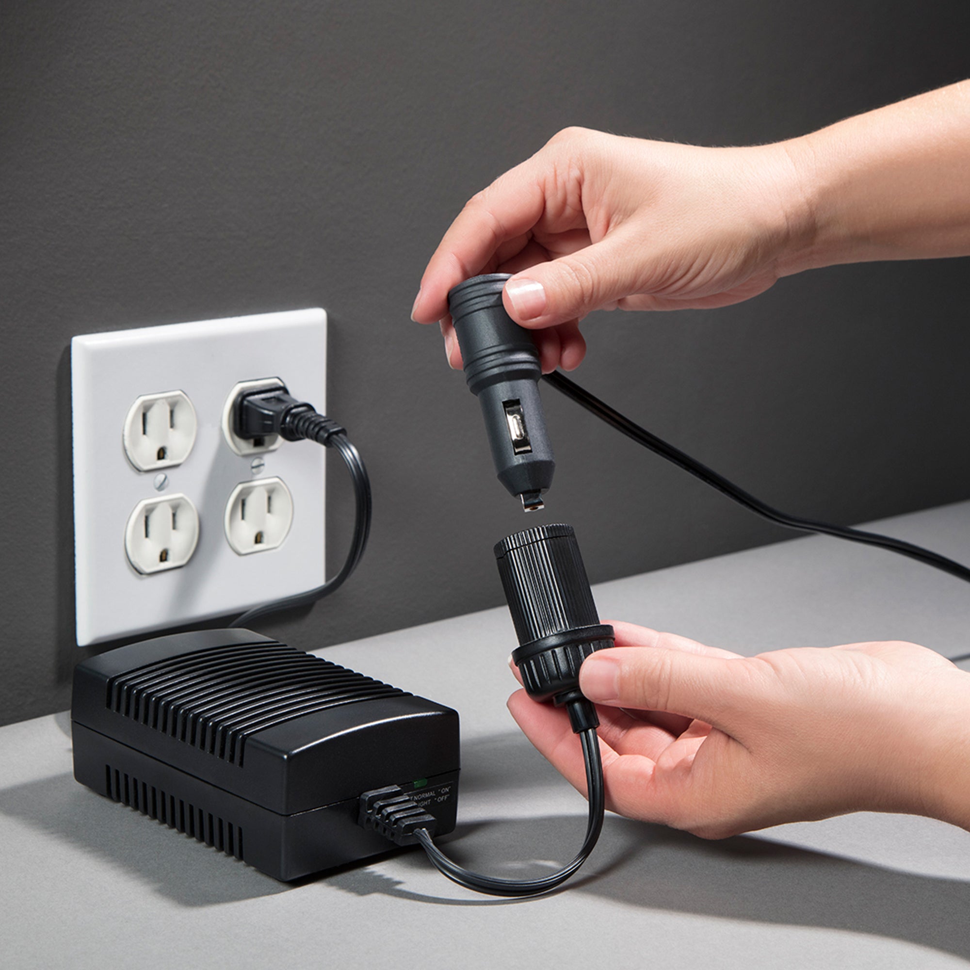 Koolatron AC to DC power adapter plugged into a white indoor AC receptacle on a dark gray wall with a person’s hands inserting a 12V plug into the DC adapter end