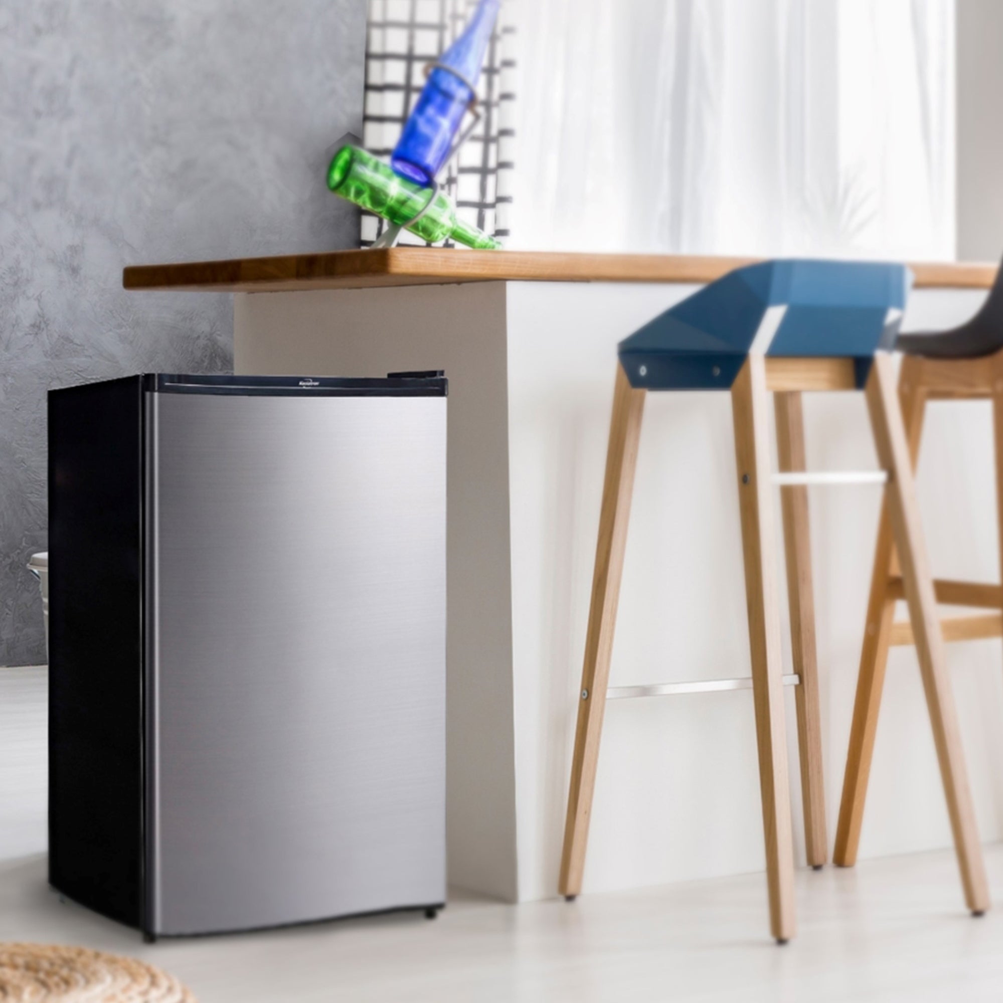 Black and stainless steel compact fridge with freezer beside a white breakfast bar with wooden countertop and two wood and plastic bar stools. Text above reads "Koolatron stainless steel compact fridge with freezer"