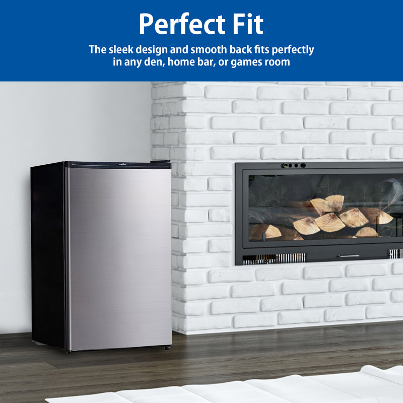Lifestyle image of black and stainless steel compact fridge with freezer on a dark gray wooden floor to the left of a white painted brick fireplace with gas insert. Text above reads, "Perfect fit: The sleek design and smooth back fits perfectly in any den, home bar, or games room"