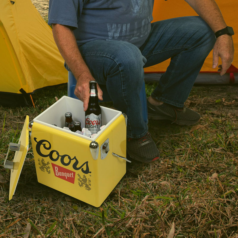 Lifestyle image of a person wearing jeans and a blue t-shirt sitting in front of two yellow dome tents and lifting a bottle of Coors Light beer out of the open ice chest