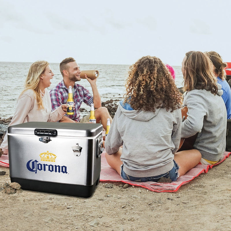 Lifestyle image of a group of people sitting on a blanket on the beach and drinking beer with the ocean in the background and a Corona 54 quart ice chest, closed, in the foreground