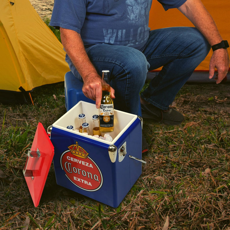 Lifestyle image of a person wearing jeans and a blue t-shirt sitting in front of two yellow dome tents and lifting a bottle of Corona beer out of the open ice chest