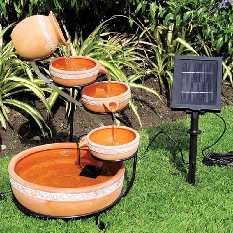 Lifestyle image of cascading terracotta solar fountain and solar panel set up on green grass with a garden plot behind