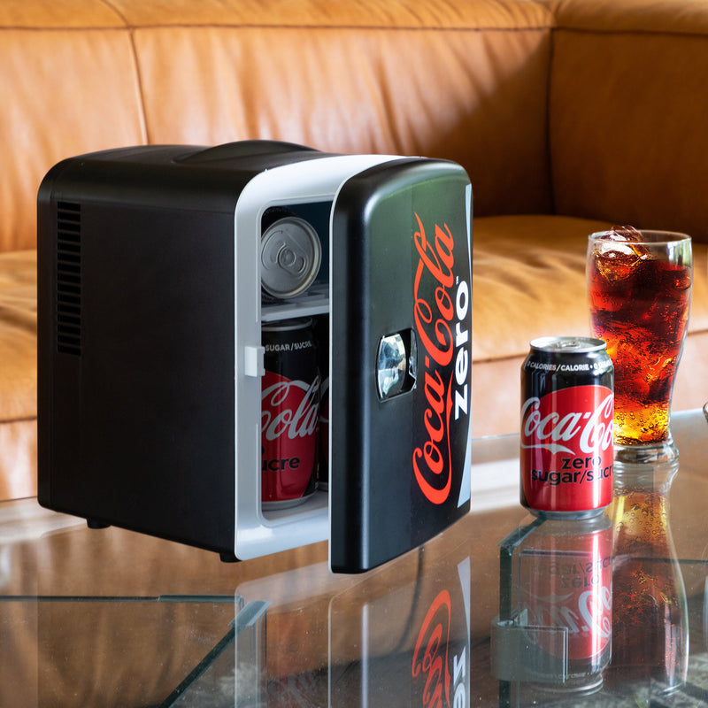 Lifestyle image of Coca-Cola Coke Zero 6 can mini fridge, partly open with cans inside, on a glass coffee table with a tan leather sofa behind it and a can and glass of Coke Zero to the right