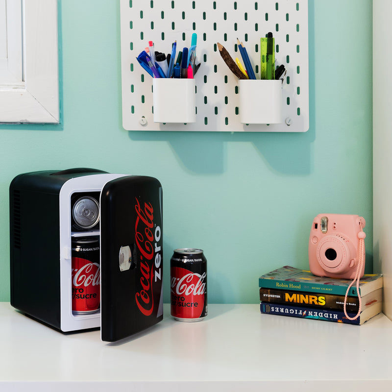 Lifestyle image of Coca-Cola Coke Zero 6 can mini fridge, partly open with cans inside, on a white desktop with an aqua wall behind. There is a can of Coke Zero, a stack of books, and a pink camera to the right of the fridge and two cups of pens and pencils attached to the wall above
