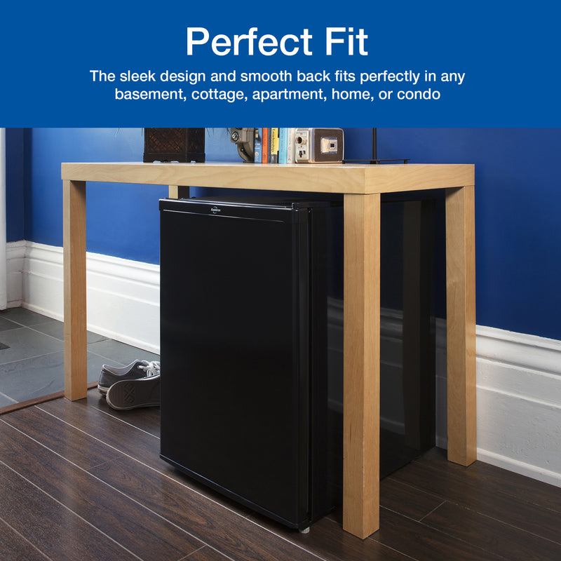 Lifestyle image of black compact fridge with freezer under a light-colored wooden console table with dark wooden floor underneath and dark blue wall and white baseboard. Text above reads, "Perfect fit: The sleek design and smooth back fits perfectly in any basement, cottage, apartment, home, or condo"