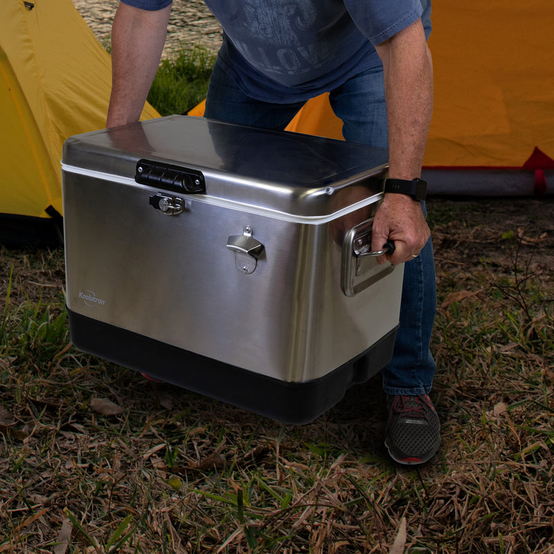 Lifestyle image of a person wearing jeans and a blue t-shirt crouching to pick up the Koolatron stainless steel 54 qt ice chest in front of two yellow dome tents