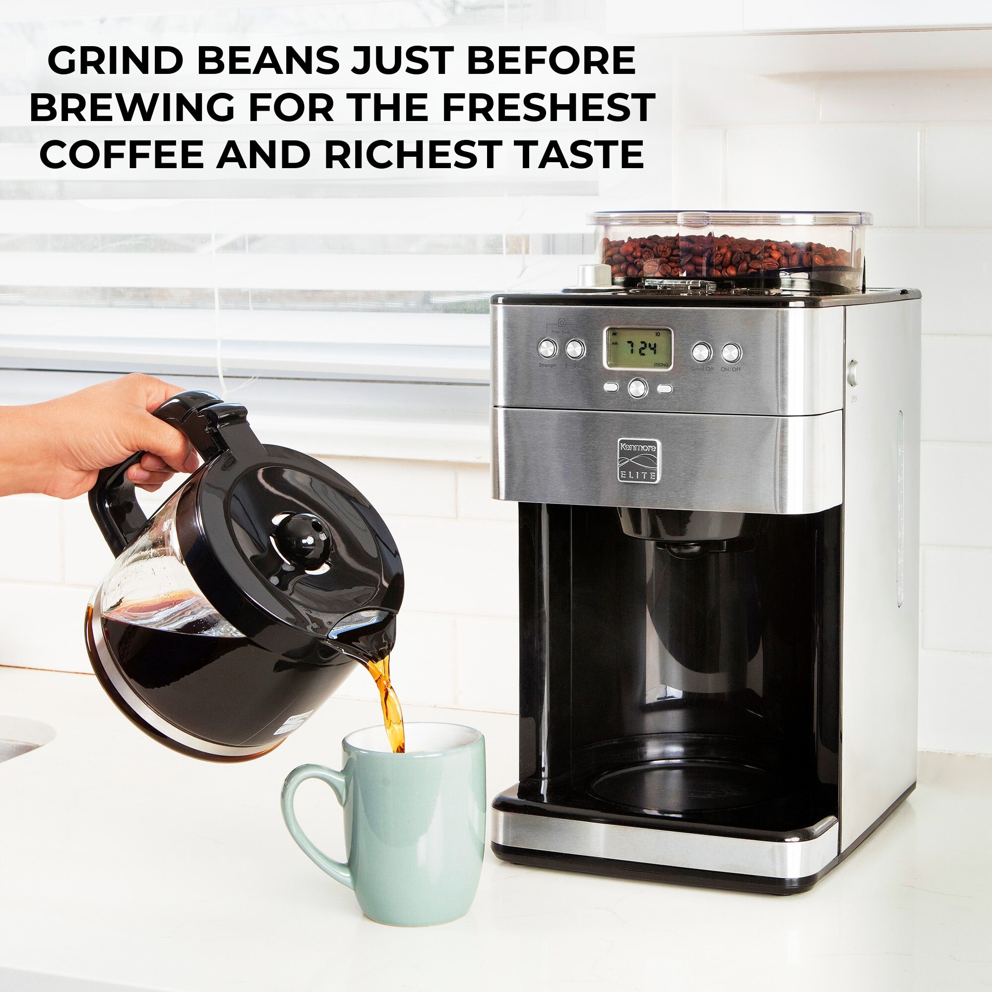 Kenmore grind and brew coffee maker on a white counter with white tile backsplash and a window with white blinds behind it and a person's hand pouring coffee from the glass carafe into a light blue mug in front. Text above reads, "Grind beans just before brewing for the freshest coffee and richest taste"