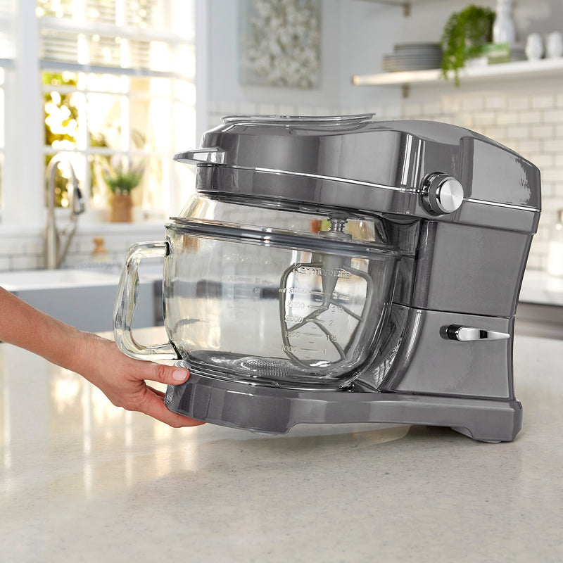 Lifestyle image of hand tilting gray Elite Ovation stand mixer to move it using the tilt-and-glide feature across a light gray kitchen counter with gray cupboards in the background