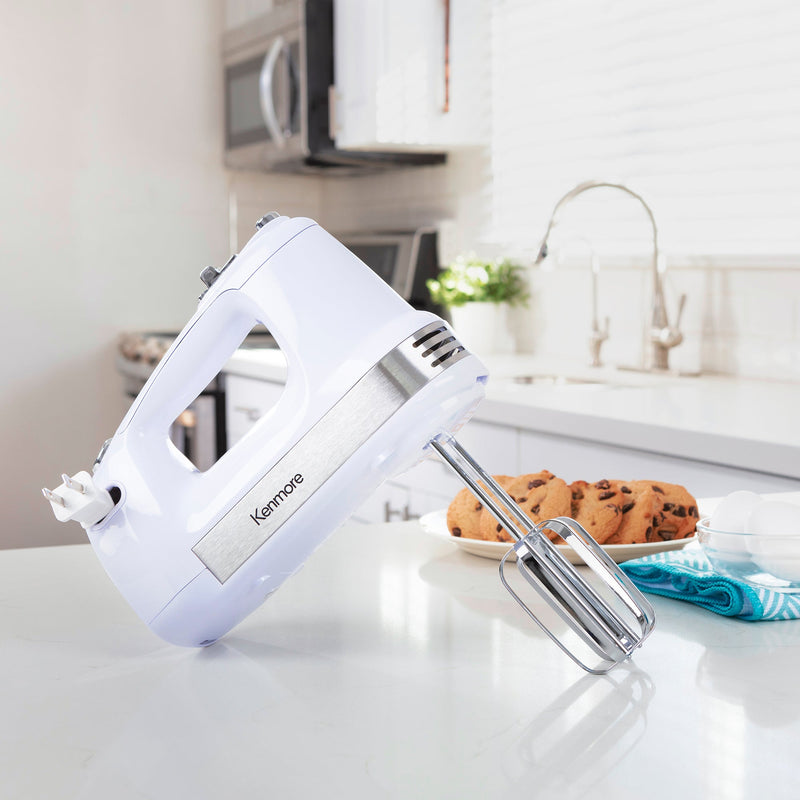 Lifestyle image of hand mixer with beaters standing on a light grey countertop with a plate of chocolate chip cookies, a turquoise dish towel, and a glass bowl of eggs beside it and a sink, oven, and microwave in the background
