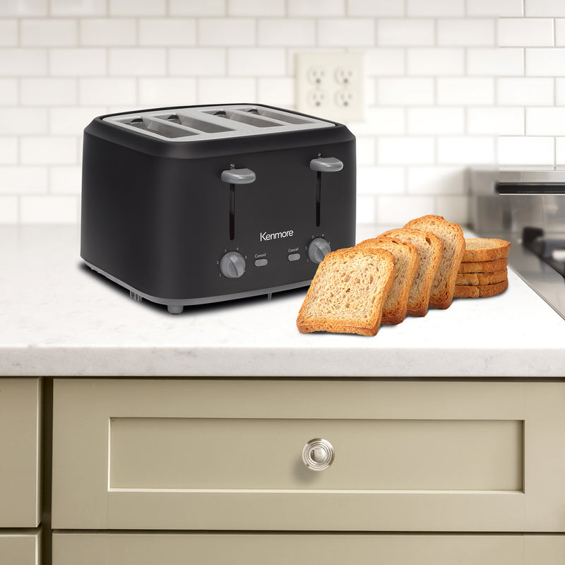 Kenmore 4-slice matte black toaster on light gray countertop over a beige drawer with a white tiled backsplash behind and slices of bread in front