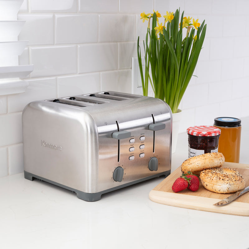 Lifestyle image of Kenmore 4-slice stainless steel toaster on light gray countertop with white tile backsplash and a pot of yellow daffodils behind. On the right is a wooden cutting board with strawberries, uncut bagels, knife, and two jam jars