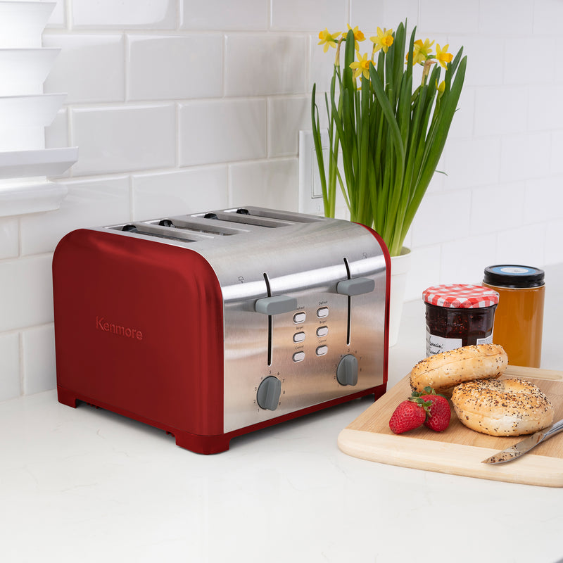 Lifestyle image of Kenmore 4-slice red stainless steel toaster on light gray countertop with white tile backsplash and a pot of yellow daffodils behind. On the right is a wooden cutting board with strawberries, uncut bagels, knife, and two jam jars