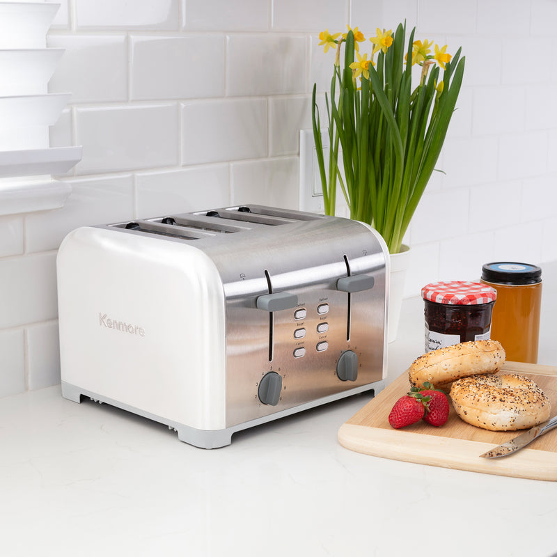 Lifestyle image of Kenmore 4-slice white stainless steel toaster on light gray countertop with white tile backsplash and a pot of yellow daffodils behind. On the right is a wooden cutting board with strawberries, uncut bagels, knife, and two jam jars