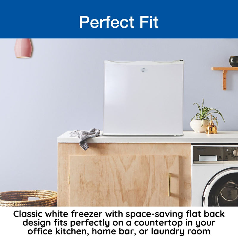 Lifestyle image of closed mini freezer on a white countertop with a light-colored wooden cupboard and a front-load washing machine below, a light gray wall behind, and a washcloth, woven basket, and potted plant to the left and right. Text above reads, "Perfect fit," and text below reads, "Classic white freezer with space-saving flat back fits perfectly on a countertop in your office kitchen, home bar, or laundry room"