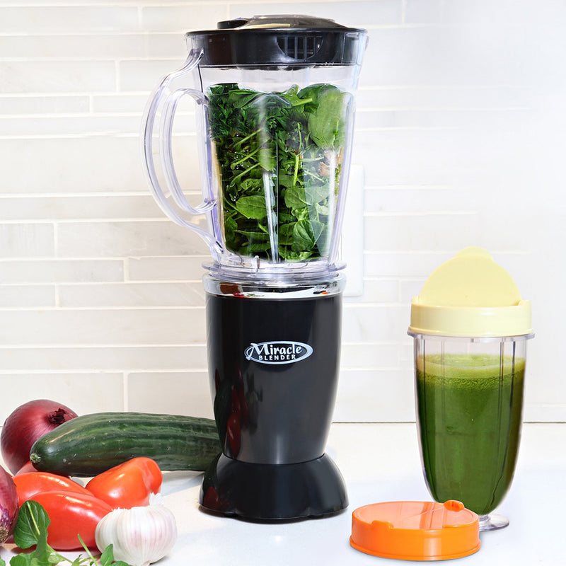 Lifestyle image of blender on a white countertop with pitcher filled with green vegetables, one personal cup with lid filled with green smoothie, and vegetables beside