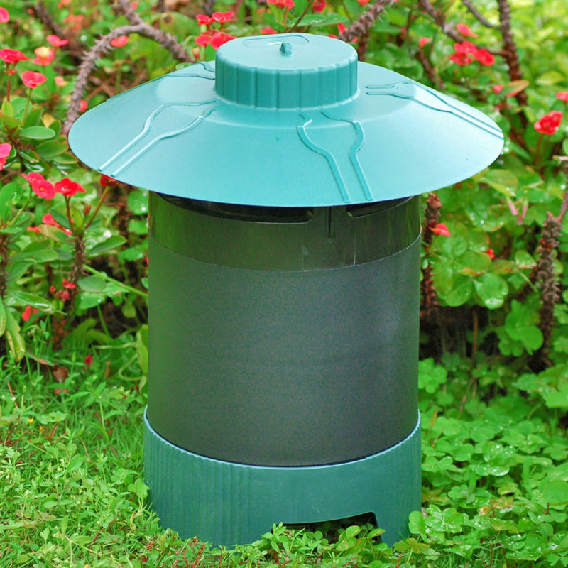 Lifestyle image of Bite Shield Protector 1/4 acre mosquito trap placed outdoors on the ground with green grass and clover under it and a plant with red stems, bright red flowers, and green leaves behind