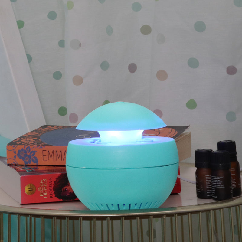 Lifestyle image of aqua indoor insect trap with nightlight turned on placed on a bedside table with two books stacked behind it and three brown glass essential oil bottles to the right. There is a white curtain with colored polkadots in the background.