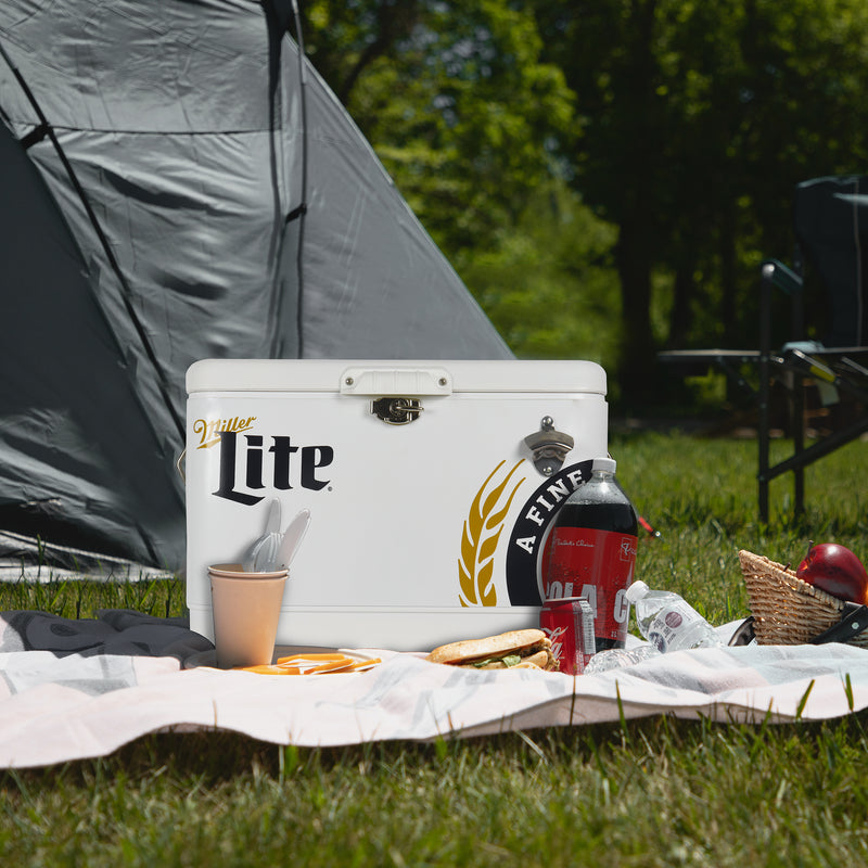 Lifestyle image of Miller Lite 54 qt ice chest on a picnic blanket on the grass with fresh fruit and a bottle and can of soda in front and a teal dome tent behind