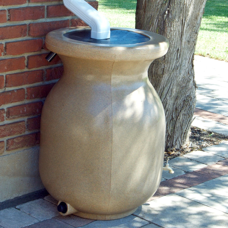 Lifestyle image of rain barrel set up on a brick pathway under a white downspout with a red brick wall, a tree, and grass in the background