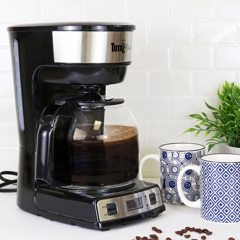 Lifestyle image of programmable coffeemaker with brewed coffee in it on a white countertop with a white tile backsplash. There is a blue and white mug to the left and a plant and wooden cutting board to the right