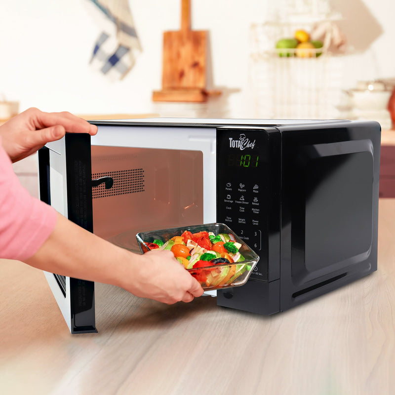 Lifestyle image of microwave on a light-colored wooden counter with a person’s hand placing a glass dish of mixed vegetables inside