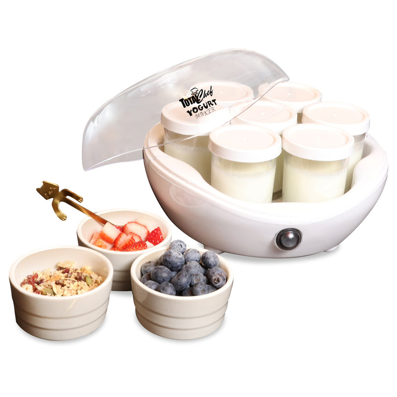 Product shot on white background of yogurt maker with lid half off and jars inside filled with plain yogurt and three small white bowls beside containing granola, strawberries, and blueberries. There is a gold-coloured spoon with a handle shaped like a cat in the strawberry bowl
