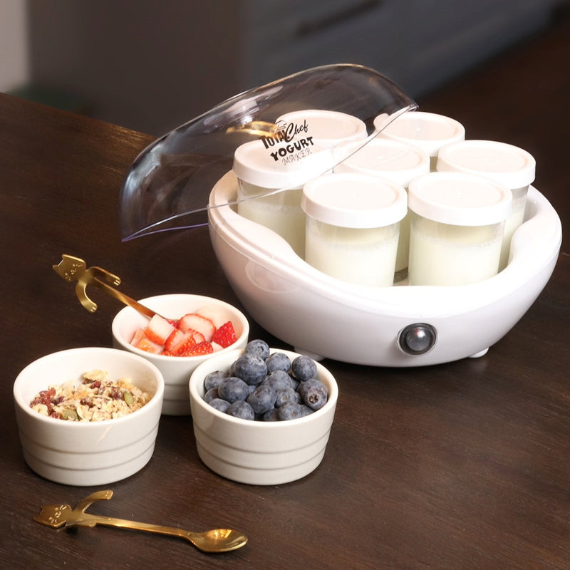 Lifestyle image of yogurt maker with lid half off and jars inside filled with plain yogurt and three small white bowls beside containing granola, strawberries, and blueberries on a dark brown wooden counter. There is a gold-coloured spoon with a handle shaped like a cat in the strawberry bowl