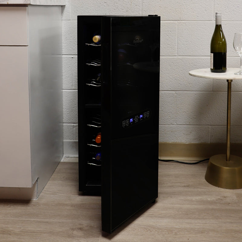 Koolatron 24 bottle dual zone wine cooler, open and filled with bottles of wine, between a white cabinet and a white and gold side table with a bottle of wine and glass on it