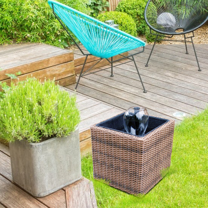 Lifestyle image of Koolscapes wicker-look pond kit with water bell set up on grass beside a wooden patio. There is a cement planter with a bright green plant on a wooden bench to the right and two deck chairs behind