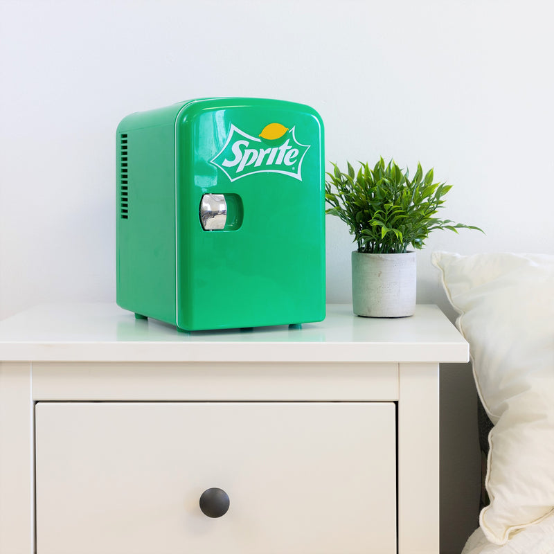 Lifestyle image of Coca-Cola Sprite 6 can mini fridge, closed, on a white bedside table with a plant in a white pot on its right and a white painted wall behind