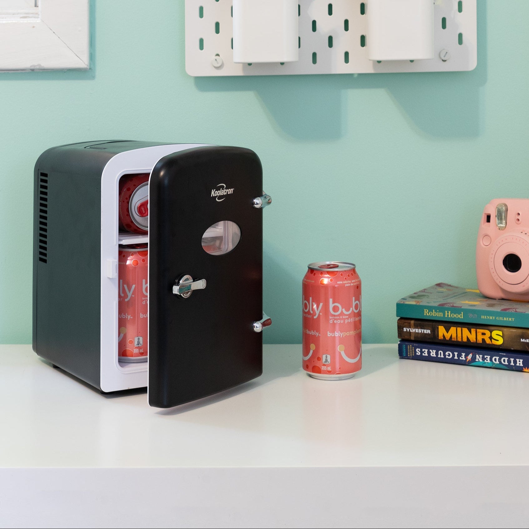 Lifestyle image of Koolatron retro 6 can mini fridge, partly open with cans inside, on a white desktop with an aqua wall behind. There is a can of soda, a stack of books, and a pink camera to the right of the fridge and two cups of pens and pencils attached to the wall above
