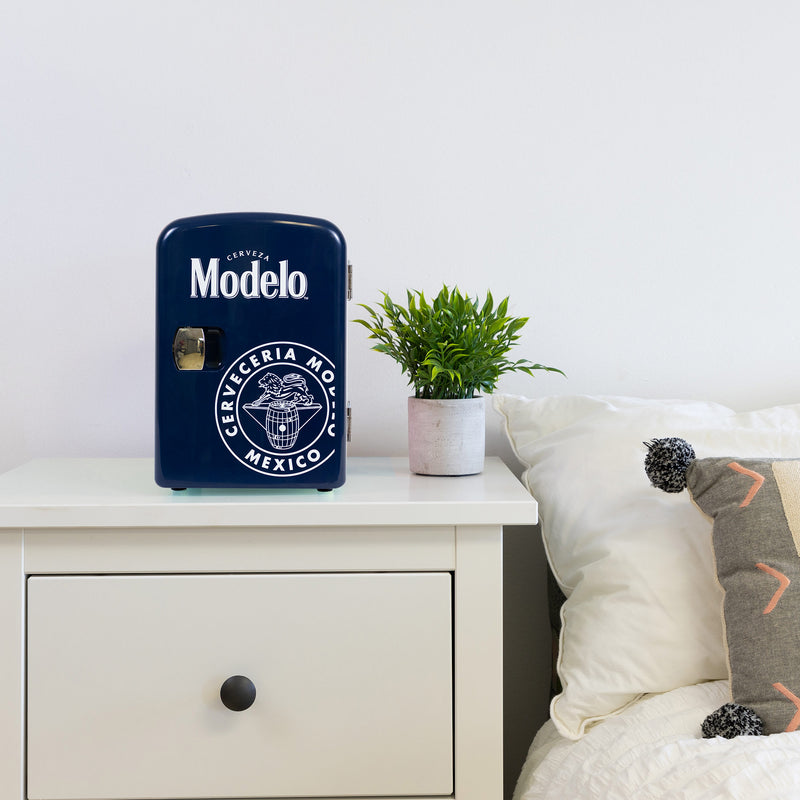 Lifestyle image of Modelo portable mini fridge, closed, on a white bedside table with a plant in a white pot on its right and a white painted wall behind